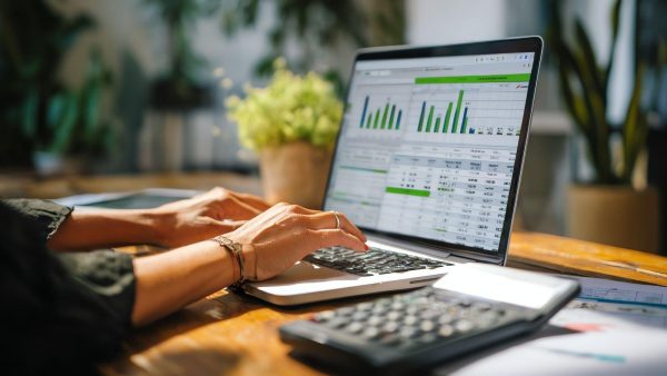 Hands typing on a laptop screen displaying green bar charts and a financial dashboard at a wooden desk.