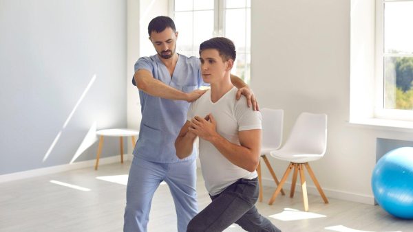 Physiotherapist guides a client through a chest-and-shoulder stretch in a bright rehab room.