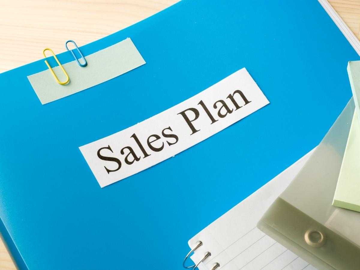 Blue binder with a white 'Sales Plan' label and colorful paperclips on a light desk, indicating planning materials.
