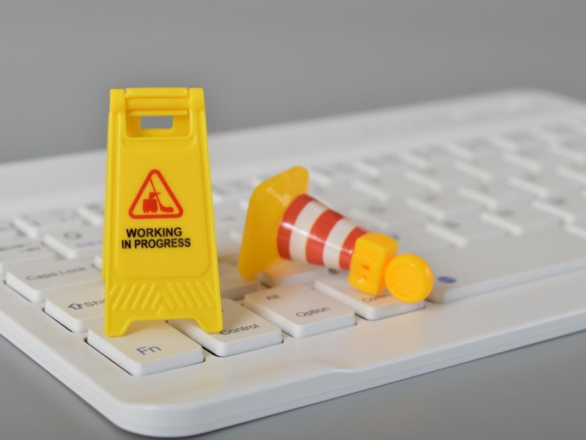 Yellow caution sign reading 'Working in Progress' and a striped traffic cone on a keyboard, signaling ongoing IT maintenance.