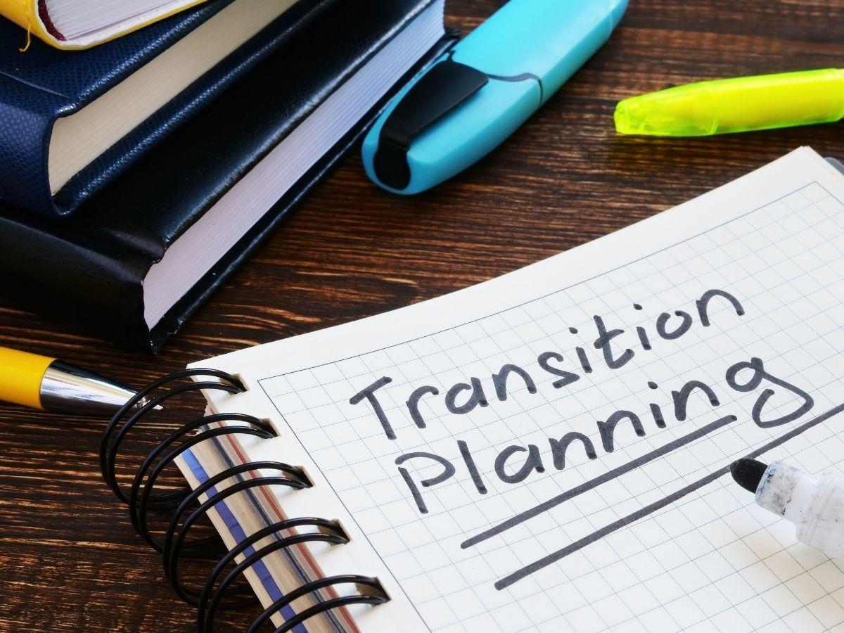 Notebook page shows the words 'Transition Planning' with a yellow pen, spiral rings, and stacked books on a wooden desk, surrounded by highlighters.