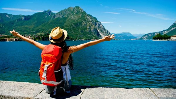 Woman with an orange backpack sits on a stone ledge by a blue lake, arms outstretched toward green mountains under a clear sky.