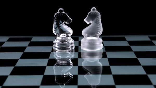 Two frosted glass knight chess pieces facing each other on a reflective checkered board.