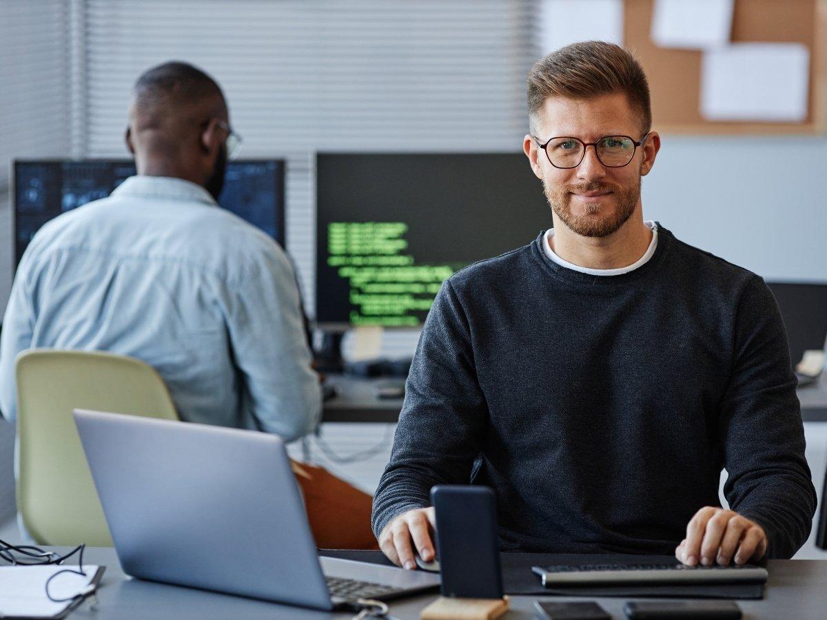 Man with glasses and beard sits at a desk with a laptop and phone, while another developer codes on green-text monitors in the background.