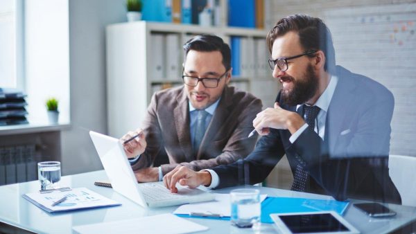 Two businessmen collaborate over a silver laptop at a glass desk in a bright office, reviewing documents together.