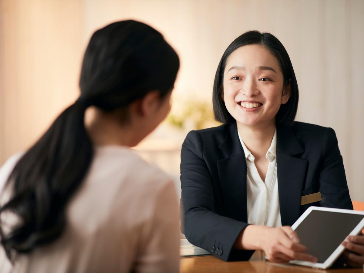 Smiling woman in a black blazer shows a tablet to a seated colleague during a business meeting in a bright office.