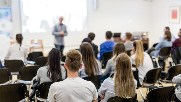 Speaker presenting to a classroom full of students seated in rows, facing a projector screen at the front of the room.
