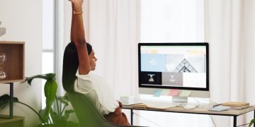 Woman at a desk stretches upward with arms raised, seated in a bright office in front of a computer monitor.