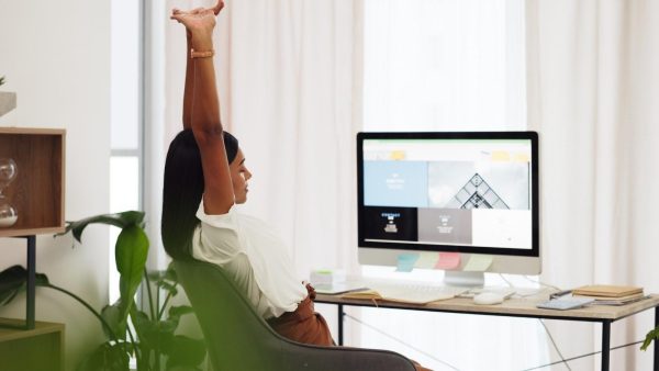 Woman at her desk stretching with arms raised, in a bright home office with a computer monitor and plants nearby.