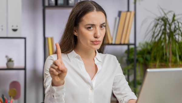 Professional woman in a white blouse sits at a laptop, raising her index finger to make a point in a bright office.