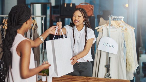 Smiling sales associate accepts a shopping bag from a customer in a clothing boutique; racks of clothes and a 'SALE' sign are visible in the background.