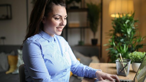 Woman smiling while typing on a laptop at a wooden desk in a cozy home office with plants nearby.