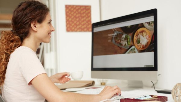 Side profile of a woman with curly hair sitting at a desk, typing on a large monitor while holding a white cup in a bright home office.