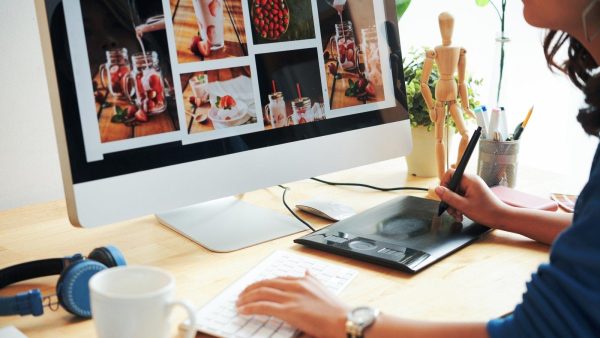 Person editing a photo collage on a large monitor using a pen tablet at a desk with headphones and a mug nearby.