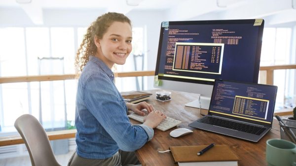 Smiling woman in a denim shirt typing at a wooden desk with a large monitor and a laptop displaying code.