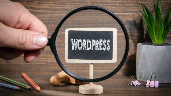 Hand holds a magnifying glass over a small chalkboard sign that says WORDPRESS on a wooden desk.
